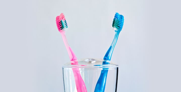 Photo of two tooth brushes, one pink, one blue, in a glass on a light grey background
