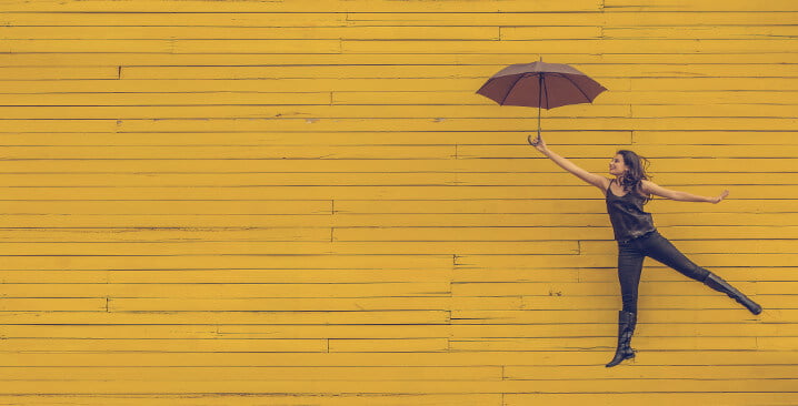 Photo of a woman jumping with an umbrella in front of a yellow brick wall