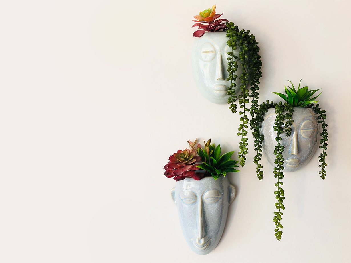 Three face-shaped wall mounted planters with succulents growing out of the top, on a beige background.