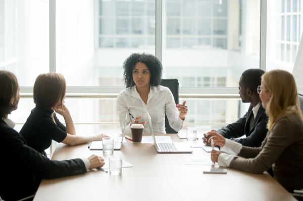 People around a table listening to a woman speaking