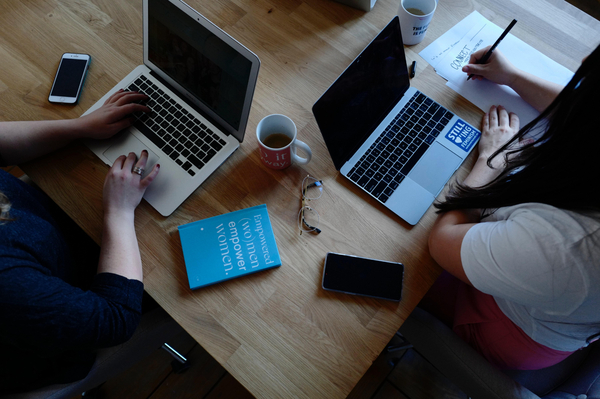 Two people working in front their laptops, cups of coffee and notebooks on the table as well