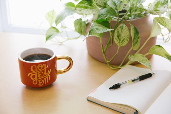 a cup of tea or coffee next to a plant, a pen and pad of paper