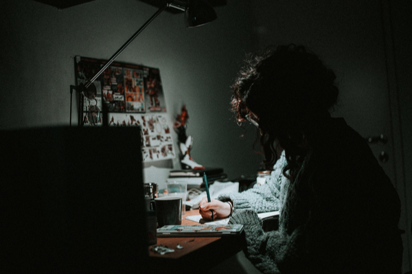 a person writing at a lit desk in a darkened room