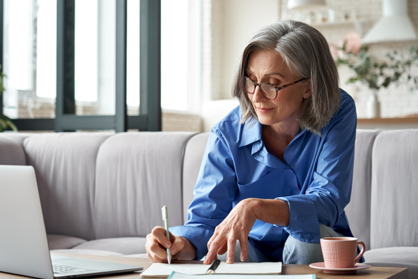 A woman sat on a sofa working from a coffee table on a laptop and making notes in a notepad.