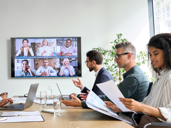 A hybrid business meeting with some participants sat at a table with others on a large screen