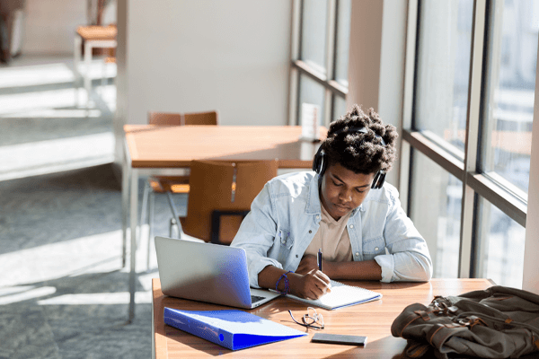 Student sat at a desk with headphones in whilst writing in a notebook. Kooth