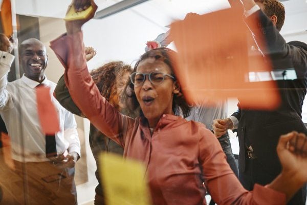 Diverse group of ecstatic businesspeople cheering together over a winning idea while brainstorming with sticky notes on a glass wall in a modern office