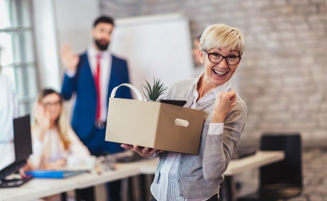 A mature woman smiling and holding a cardboard box of belongings as coworkers wave goodbye