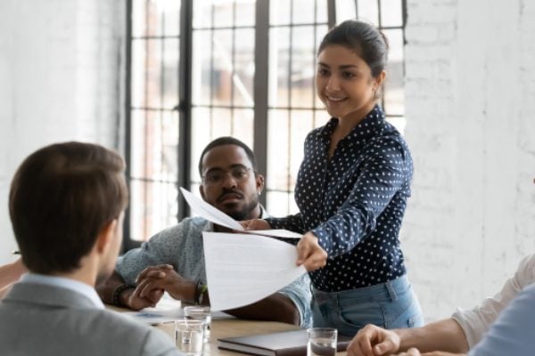 A smiling young employee hands a document to a colleague