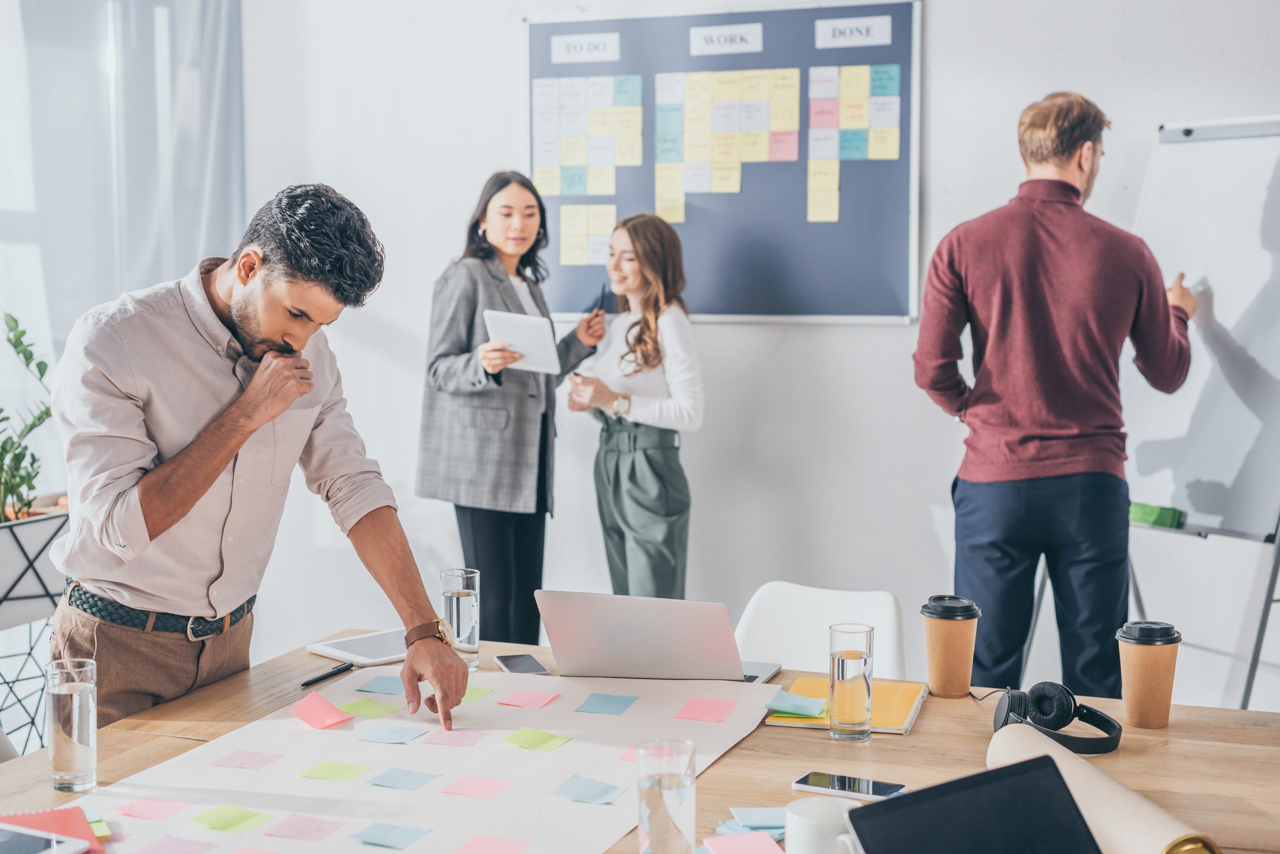 selective focus of pensive mixed race scrum master pointing with finger at sticky note near coworkers