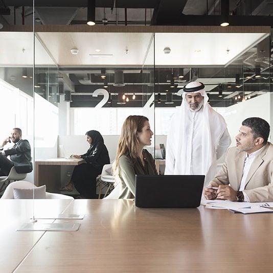 A photo of multi-ethnic Arab, Caucasian and Middle Eastern business people discussing at conference table in modern office. Professionals are with laptop. Some are in formals and others are wearing traditional attire. Dubai, United Arab Emirates.