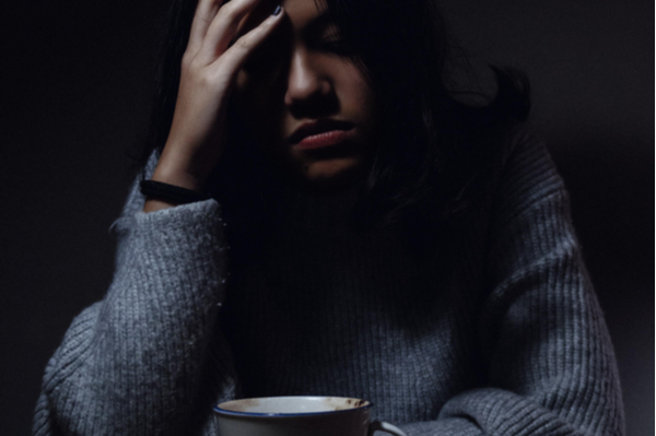 A sad looking lady with her eyes shut, a cup of coffee and her head in her hands