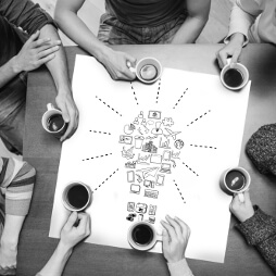 Shows peoples arms, holding cups of hot drinks, around a table, bird-eye view.