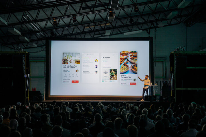 Photo of presenter in front of an audience, next to a screen